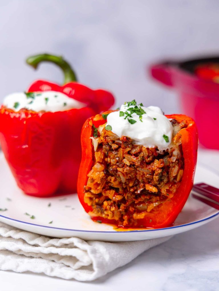 Cut-open high-protein stuffed pepper filled with ground beef and rice, topped with a dollop of greek yogurt and chopped parsley, with a second whole stuffed pepper on a white plate behind it.