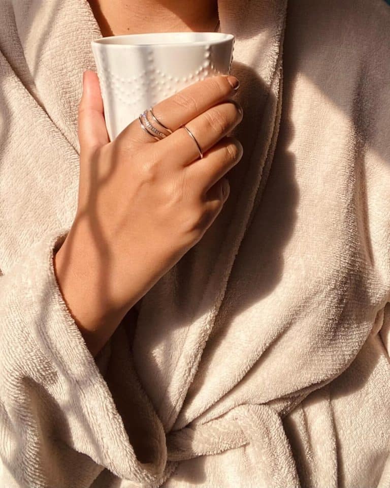 “Cozy self care morning with a woman in a soft beige robe holding a white mug of coffee or tea in warm sunlight.