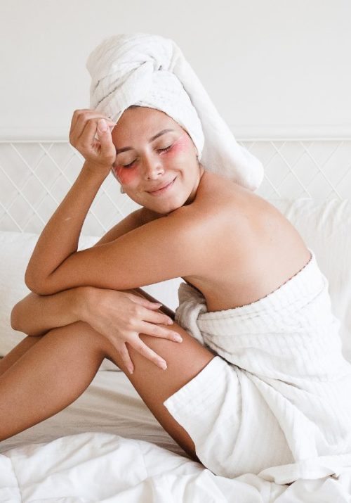 Smiling woman practicing emotional self-care in a towel, sitting on a bed with rosy cheeks, embodying relaxation and emotional wellness.