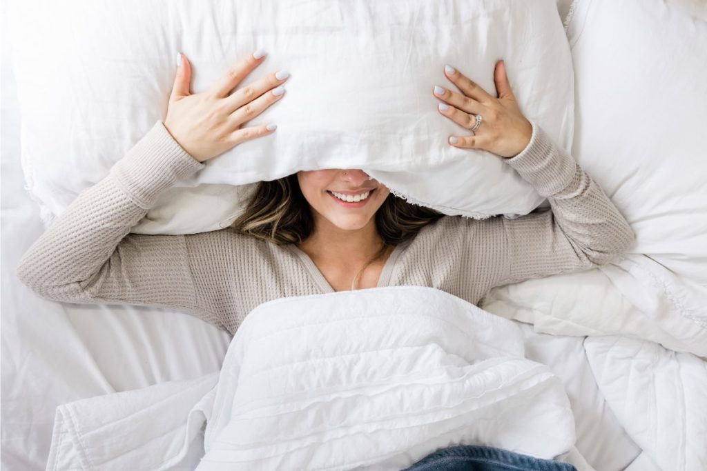 Smiling woman under a pillow, preparing to sleep deeply in a cozy bed.