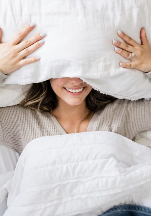 Smiling woman under a pillow, preparing to sleep deeply in a cozy bed.