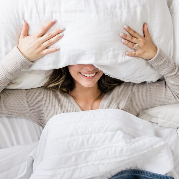 Smiling woman under a pillow, preparing to sleep deeply in a cozy bed.