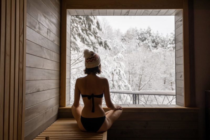 Person in a sauna during the Winter Arc, wearing a winter hat and gazing at a snowy landscape through a large window.