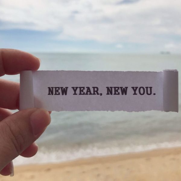 Hand holding a paper that says 'New Year, New You' on a beach, symbolizing the start of a New Years Resolution List for personal growth, self-improvement, and healthy habits.