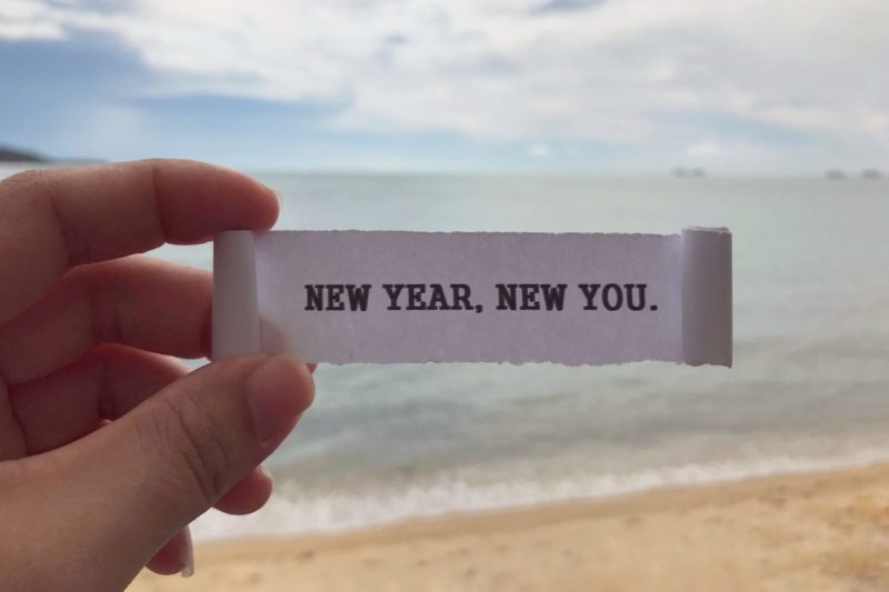 Hand holding a paper that says 'New Year, New You' on a beach, symbolizing the start of a New Years Resolution List for personal growth, self-improvement, and healthy habits.