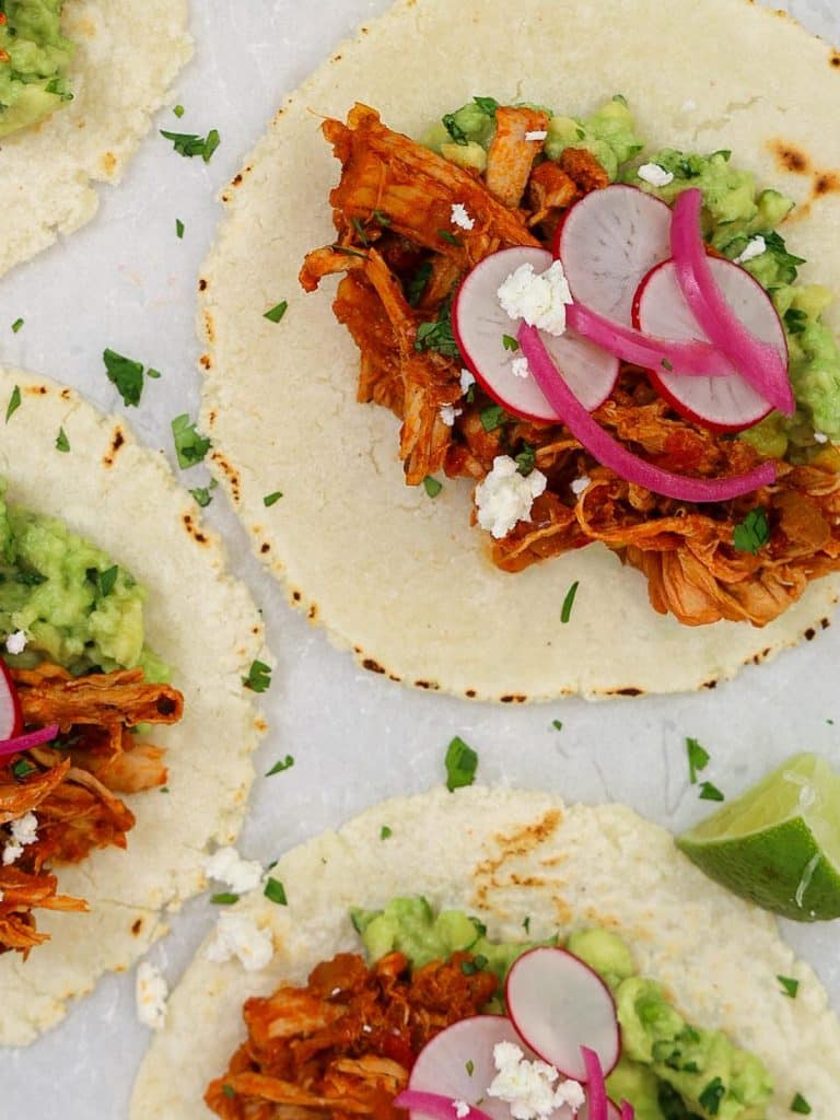 Crock pot chicken tinga recipe served on warm corn tortillas with avocado, pickled red onions, and radishes