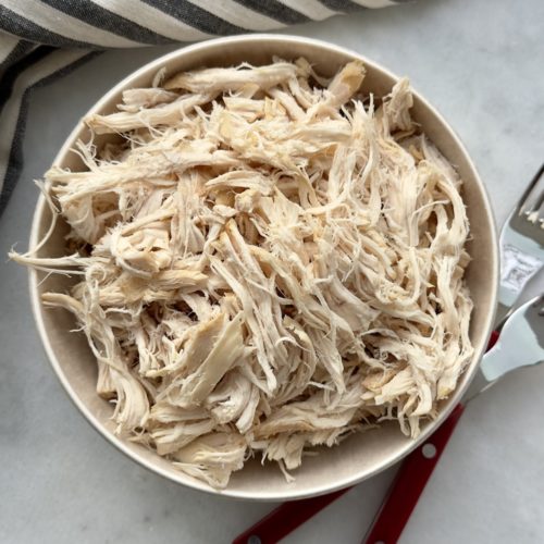 slow cooker shredded chicken in a bowl surrounded by a striped napkin and two forks used for shredding.