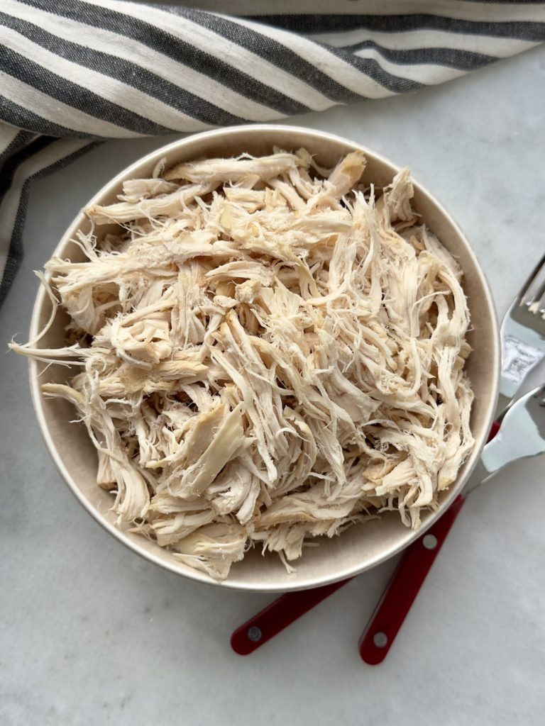 slow cooker shredded chicken in a bowl surrounded by a striped napkin and two forks used for shredding. 