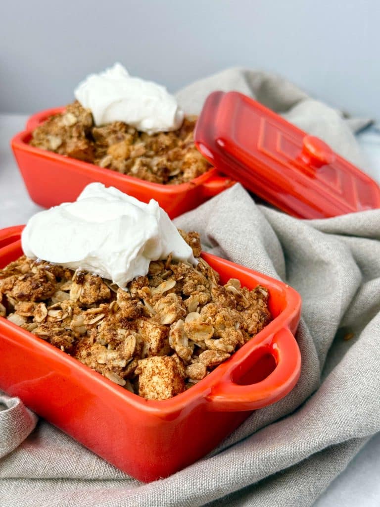 Individual red ramekins filled with high-protein apple crumble topped with Greek yogurt.