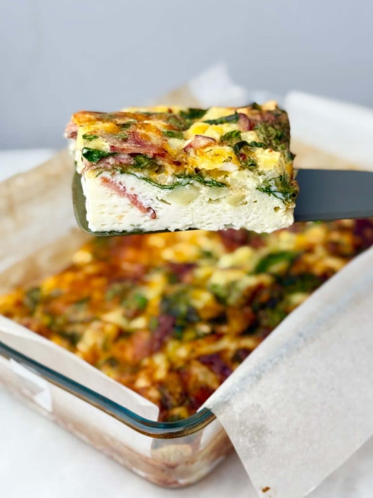 Slice of high-protein egg bake with cottage cheese, spinach, and turkey bacon lifted from a glass baking dish.