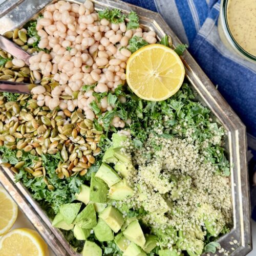 A beautifully arranged Erewhon kale white bean salad on a silver platter, featuring massaged kale, white beans, pumpkin seeds, hemp seeds, avocado chunks, and fresh lemon slices. A delicious and nutrient-rich kale and bean salad recipe.