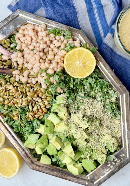 A beautifully arranged Erewhon kale white bean salad on a silver platter, featuring massaged kale, white beans, pumpkin seeds, hemp seeds, avocado chunks, and fresh lemon slices. A delicious and nutrient-rich kale and bean salad recipe.