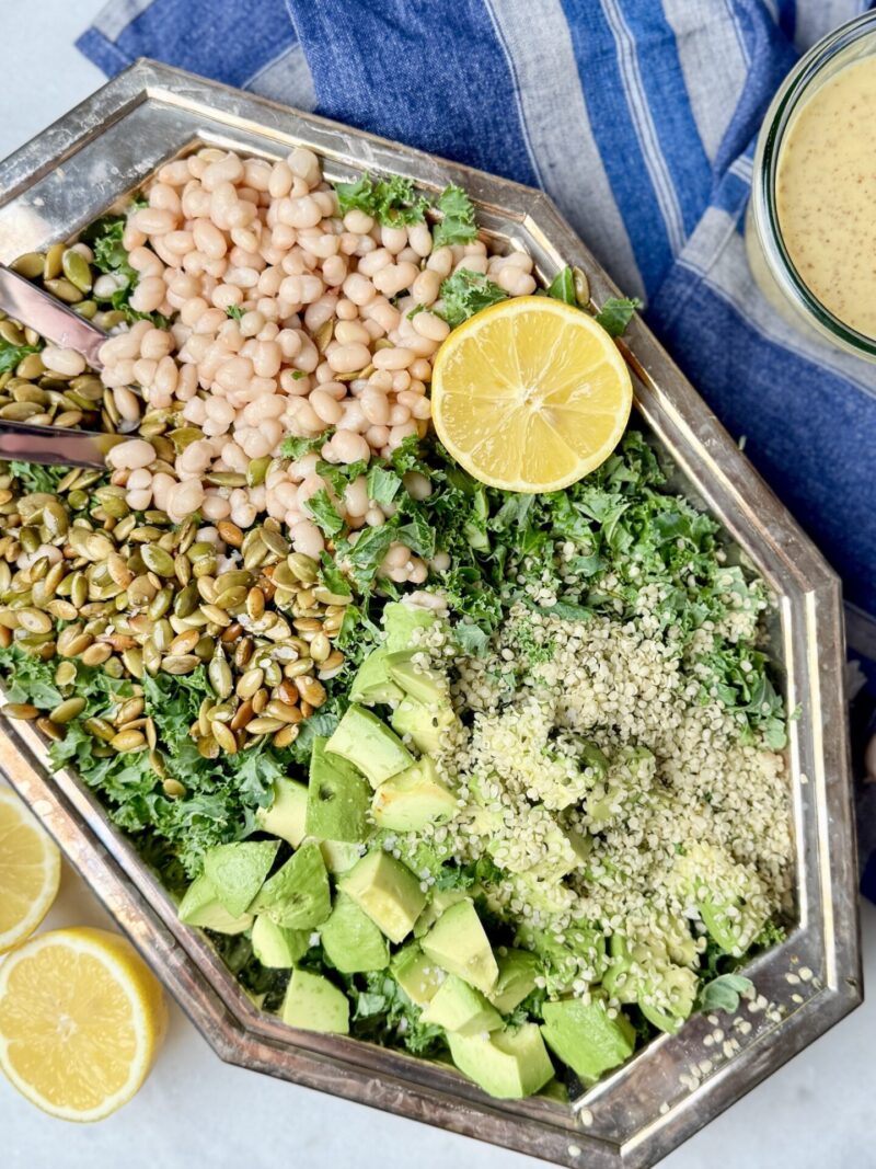 A beautifully arranged Erewhon kale white bean salad on a silver platter, featuring massaged kale, white beans, pumpkin seeds, hemp seeds, avocado chunks, and fresh lemon slices. A delicious and nutrient-rich kale and bean salad recipe.