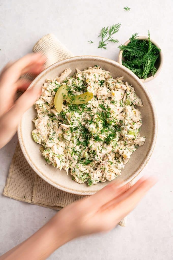 High-protein dill pickle chicken salad in a ceramic bowl garnished with fresh dill and pickle slices, with hands holding the bowl.