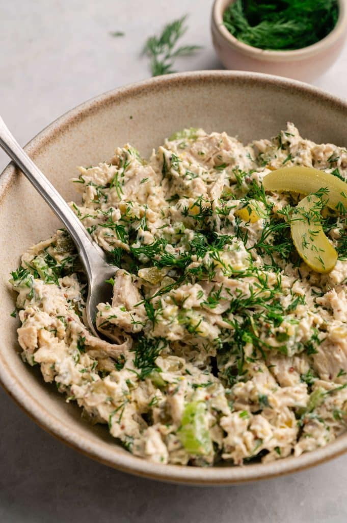 Bowl of high protein dill pickle chicken salad with shredded chicken, fresh dill, and pickle slices, served with a spoon.