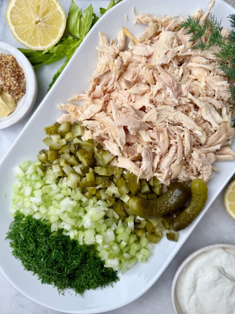 A plate of shredded chicken, diced pickles, fresh dill, and celery ready to be mixed into a dill pickle chicken salad.