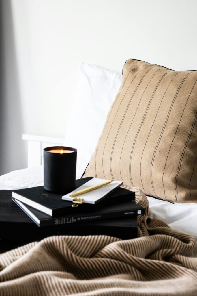 A cozy self-care setup featuring a lit candle, a notebook with a gold pen, and a warm beige pillow on a neatly made bed, aligning with a daily self care checklist.