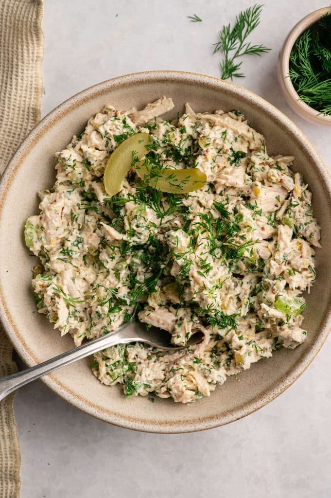 High protein dill pickle chicken salad served in a bowl with a fork, topped with fresh dill and pickle slices.