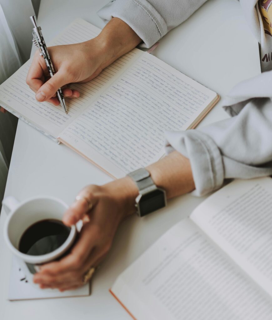 A person writing in a lined notebook with a pen while holding a cup of coffee, practicing morning pages journaling. What are morning pages? Discover the benefits of this daily writing habit.