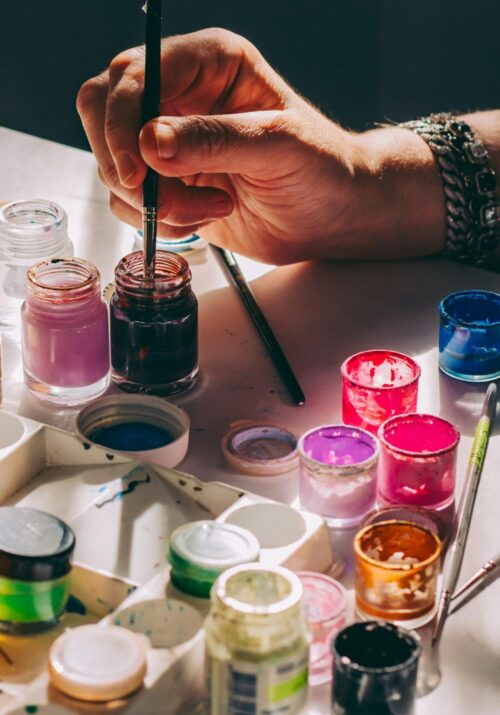 Close-up of a hand dipping a paintbrush into a jar of paint, surrounded by colorful paint pots, representing creative self-care hobbies like painting and art.