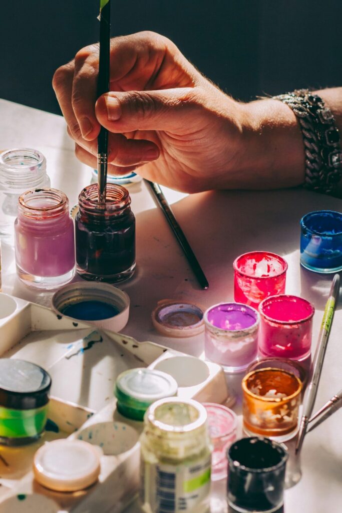 Close-up of a hand dipping a paintbrush into a jar of paint, surrounded by colorful paint pots, representing creative self-care hobbies like painting and art.