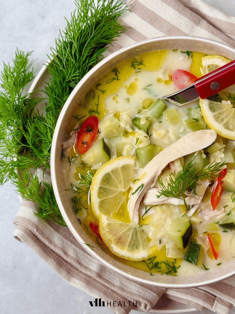 A warm and comforting high-protein low-carb chicken soup with fresh lemon slices, dill, zucchini, and orzo pasta, served in a ceramic bowl.