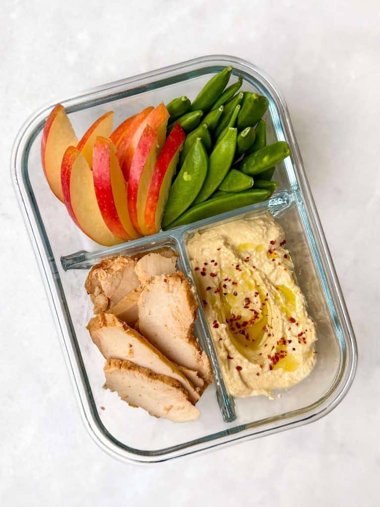 Protein lunch box ideas featuring sliced chicken breast, hummus, snap peas and apple