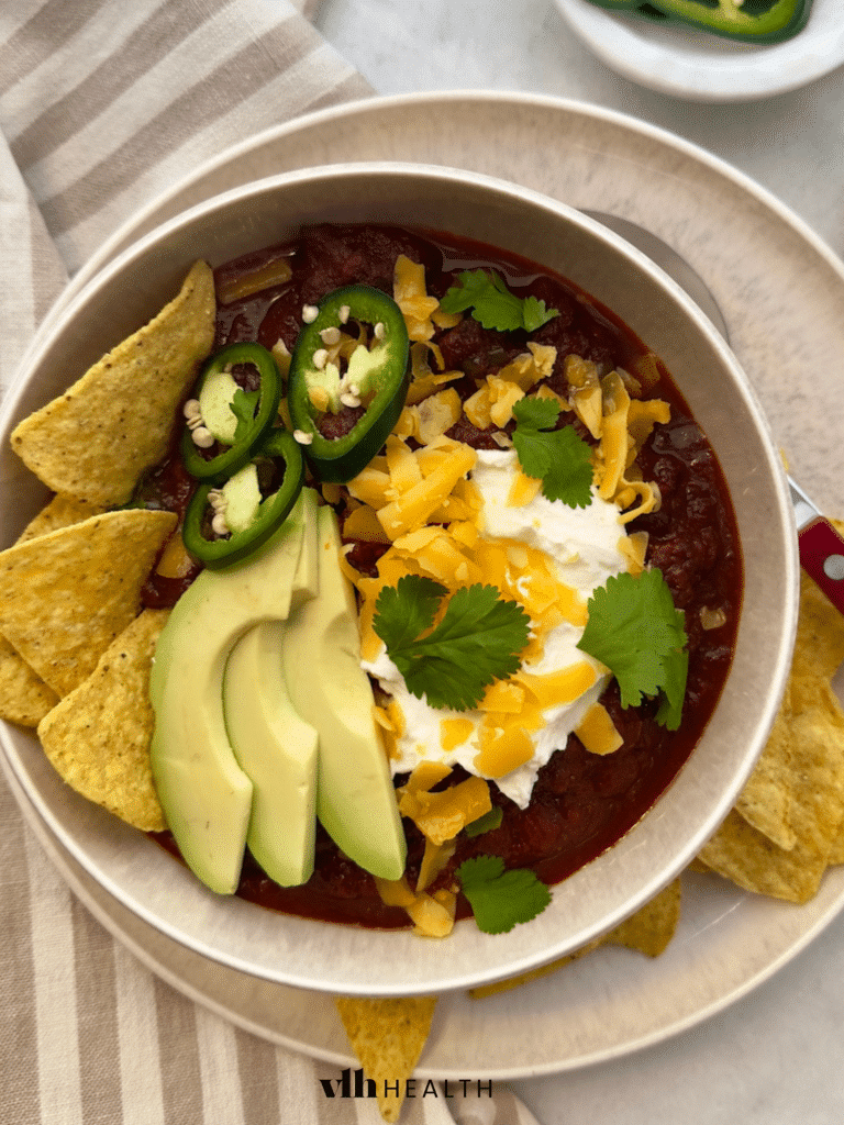 A hearty bowl of low-carb chili topped with fresh avocado slices, shredded cheddar cheese, sour cream, and jalapeños, served with tortilla chips.