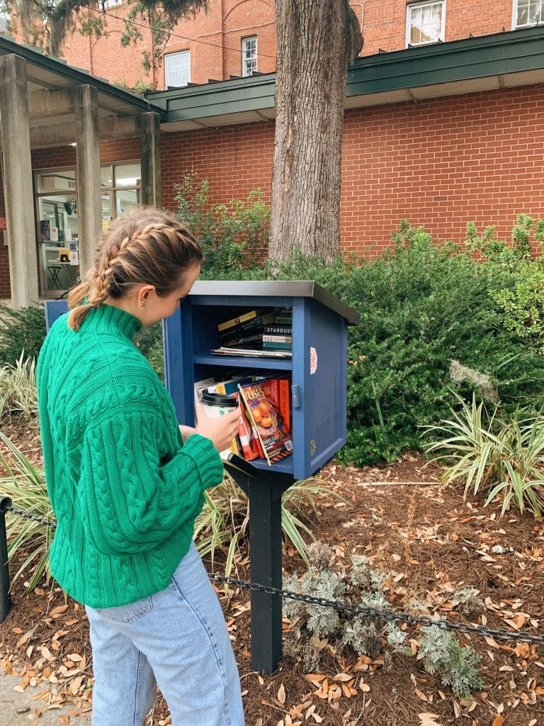 a woman in a green sweater placing books in a community library box, representing volunteering and giving back habits
