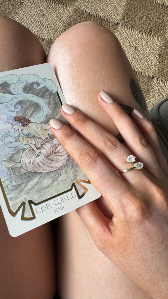 A woman holding a tarot card titled The Well with soft neutral nails and a crystal ring — a feminine hobby focused on intuition and spiritual self-care