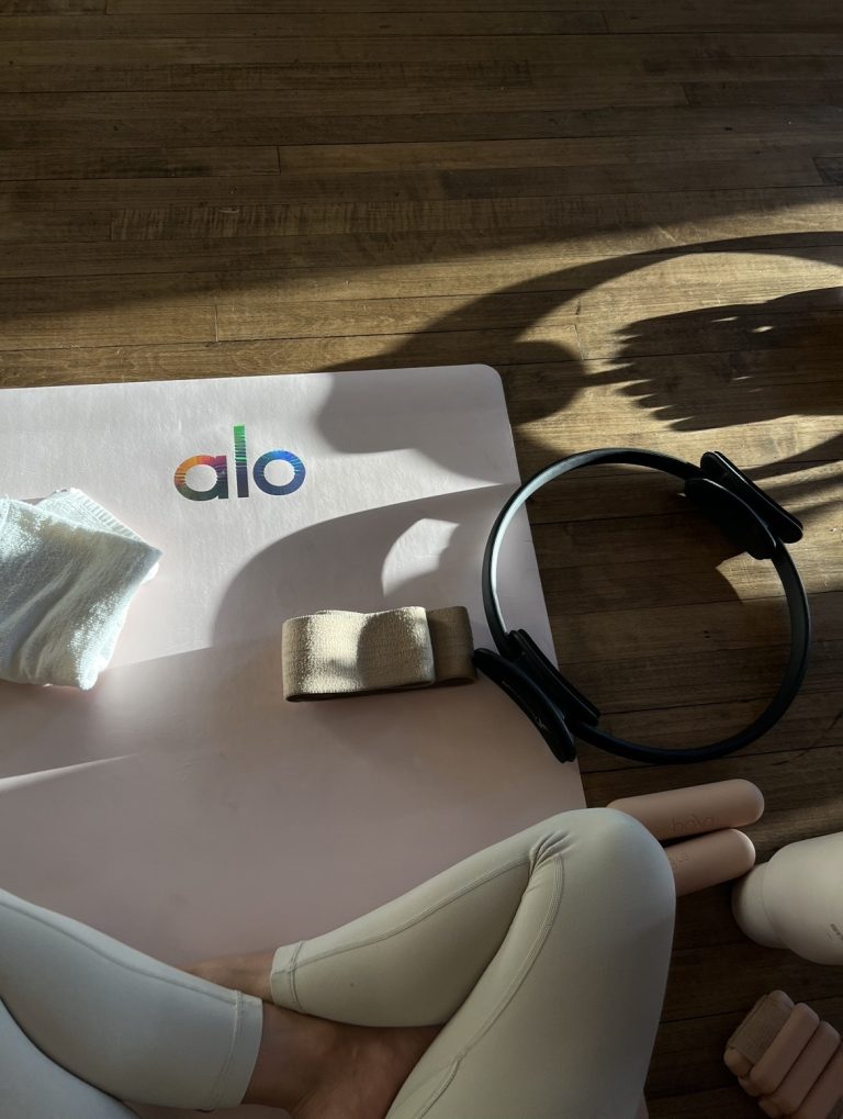 Flat lay of a Pilates arm workout setup with a pink Alo yoga mat, resistance band, Pilates ring, hand weights, and a woman in cream activewear sitting cross-legged on a wooden floor in natural sunlight.