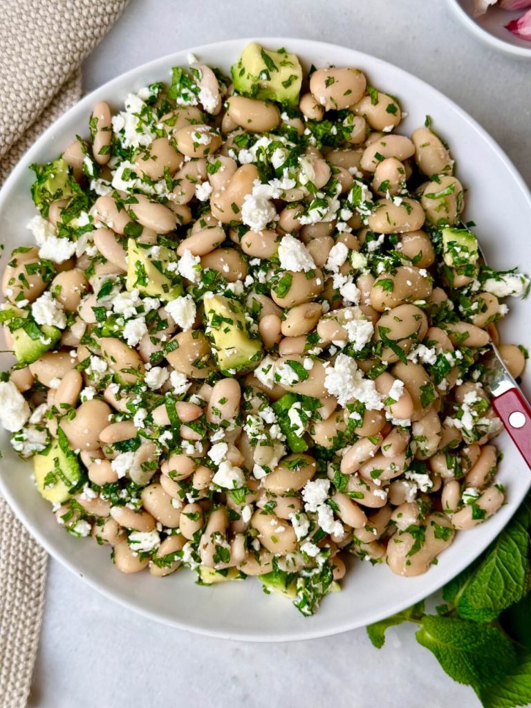 Overhead view of a Mediterranean white bean salad made with canned beans, creamy avocado, crumbled feta, and fresh chopped herbs on a white background.