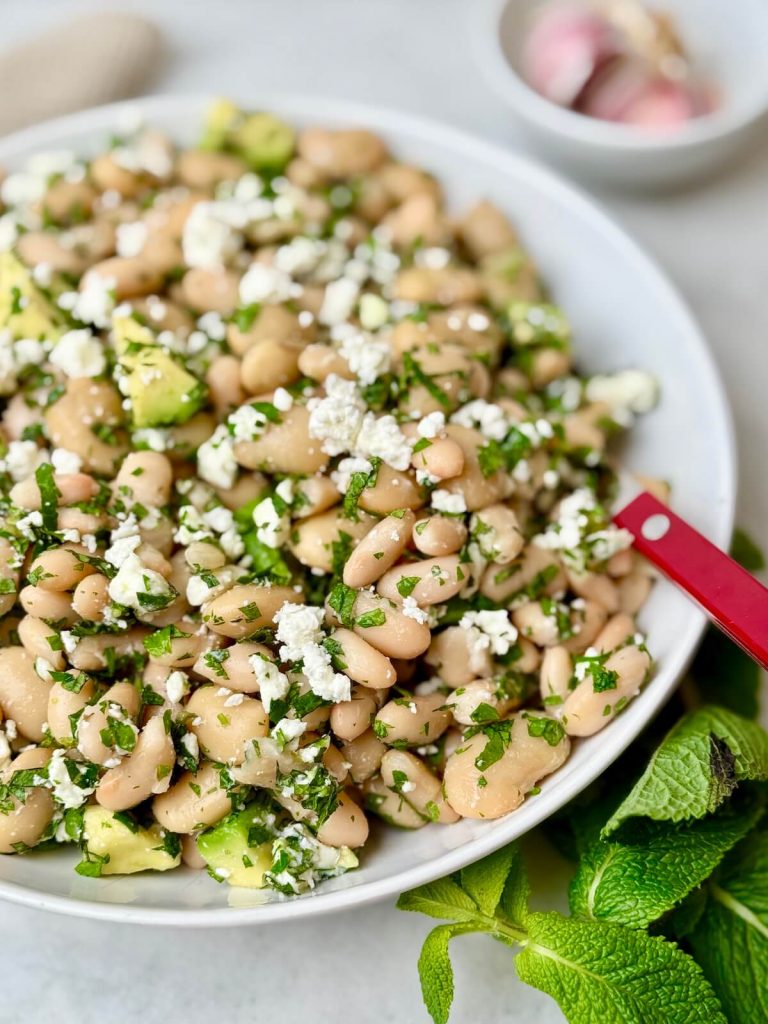 Close-up of Mediterranean white bean salad with feta cheese, avocado, and fresh herbs in a white bowl, highlighting creamy white beans and vibrant parsley.