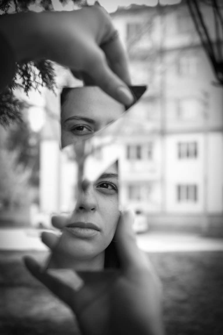 Black and white photo of a woman’s face reflected in broken mirror pieces, symbolizing self-reflection, shadow work, and emotional healing.