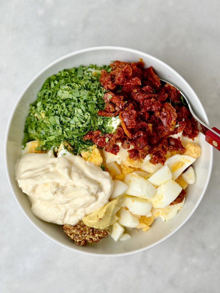 Egg salad ingredients in a bowl: chopped eggs, sun-dried tomatoes, fresh tarragon, mayonnaise, Dijon, and grainy mustard