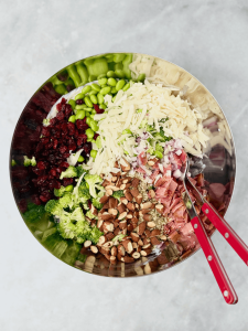 Overhead of colorful, unmixed high protein broccoli salad ingredients in a large metal bowl with red tongs, bacon, Parmesan, broccoli, edamame, and nuts.