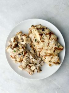Grilled and chopped chicken on a white plate, golden brown and ready to be added to the Greek chicken and orzo pasta salad for added protein and flavor.