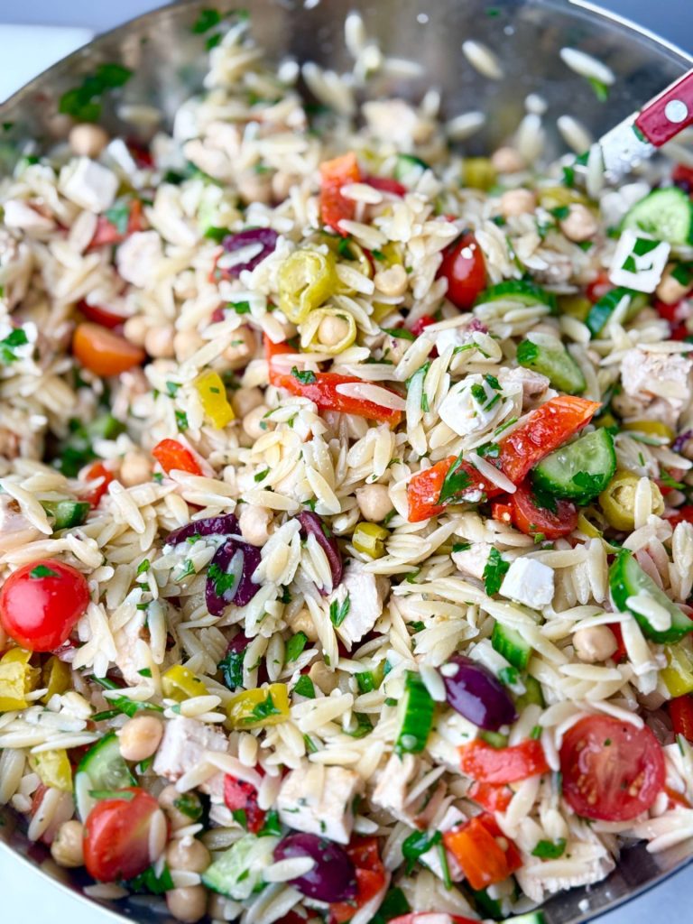 Close-up of Greek chicken and orzo pasta salad in a large stainless steel bowl, featuring orzo pasta, grilled chicken, cherry tomatoes, cucumbers, briny olives, feta, and herbs.