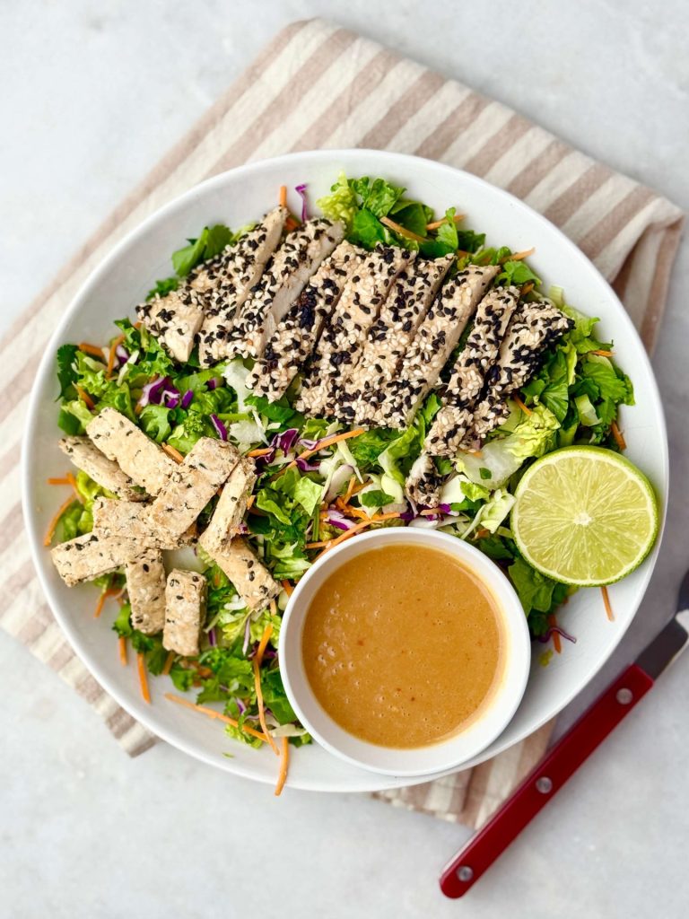 Bright top-down view of an Erewhon Asian Chicken Salad with sliced chicken breast, shredded greens, and a lime wedge beside the miso-ginger dressing.