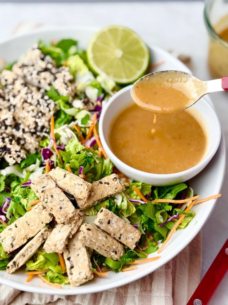 Side angle of a colorful Erewhon Asian Chicken Salad with creamy miso dressing and gluten-free sesame sticks arranged on a striped cloth.