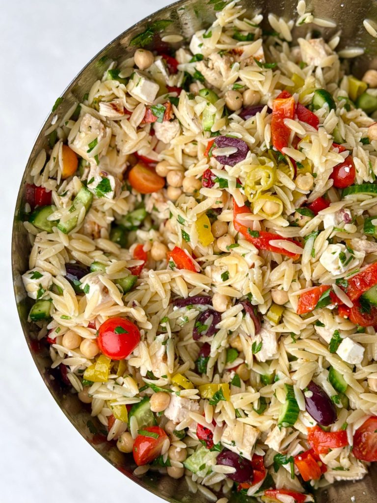 Top-down view of a vibrant Greek chicken and orzo pasta salad tossed with chickpeas, roasted red pepper, fresh parsley, grape tomatoes, and pepperoncini in a mixing bowl.