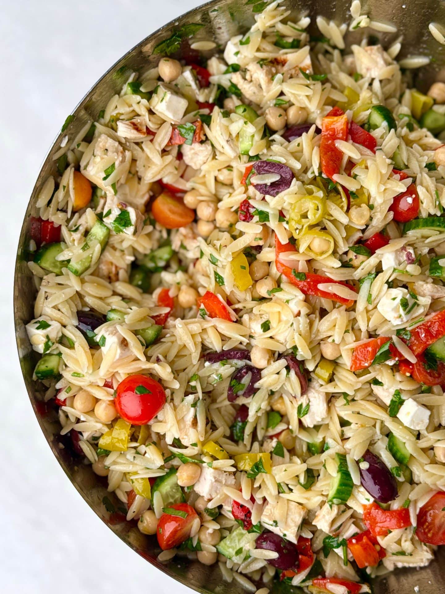 Top-down view of a vibrant Greek chicken and orzo pasta salad tossed with chickpeas, roasted red pepper, fresh parsley, grape tomatoes, and pepperoncini in a mixing bowl.