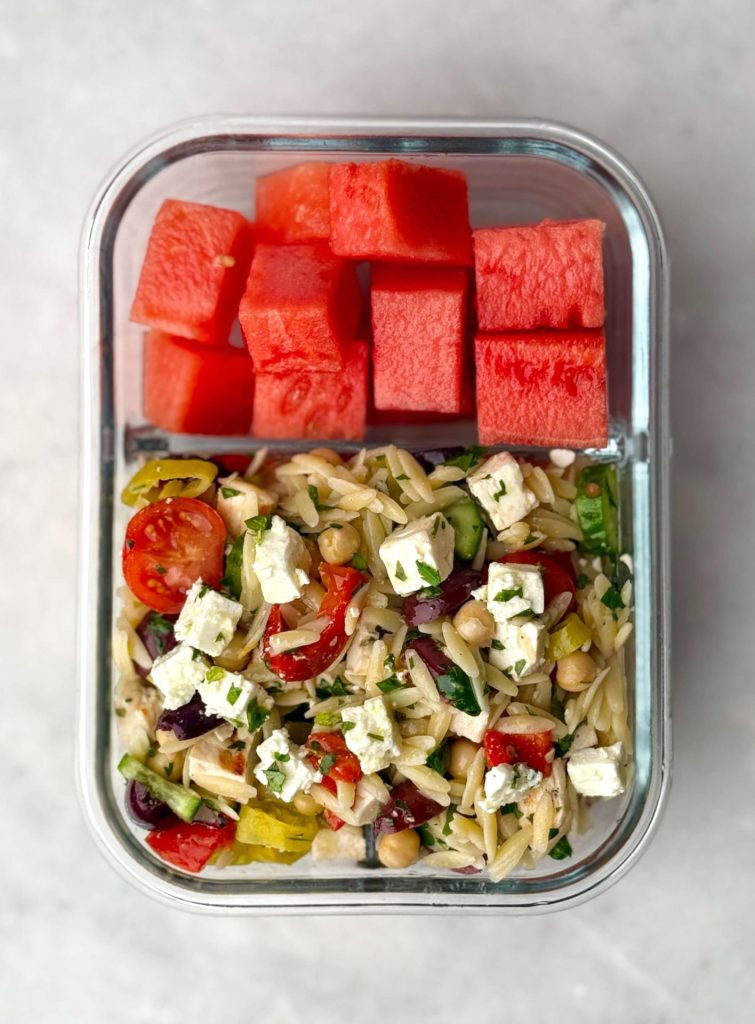 Meal prep container with Greek chicken and orzo pasta salad featuring feta cheese, cherry tomatoes, briny olives, cucumbers, and chickpeas, served with cubed watermelon. A fresh, balanced lunch idea perfect for cold pasta salads.