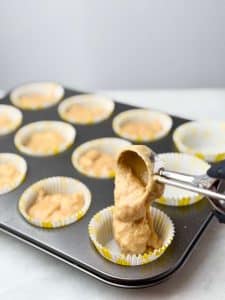 Cookie scoop filling muffin liners with high protein banana muffin batter in a metal muffin pan, ready to bake.
