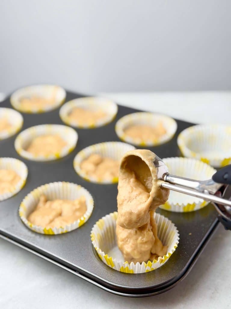 Cookie scoop filling muffin liners with high protein banana muffin batter in a metal muffin pan, ready to bake.