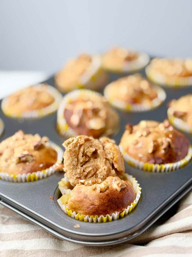 High protein banana bread muffins in a muffin pan, one muffin broken open to show soft texture and chopped walnuts on top.