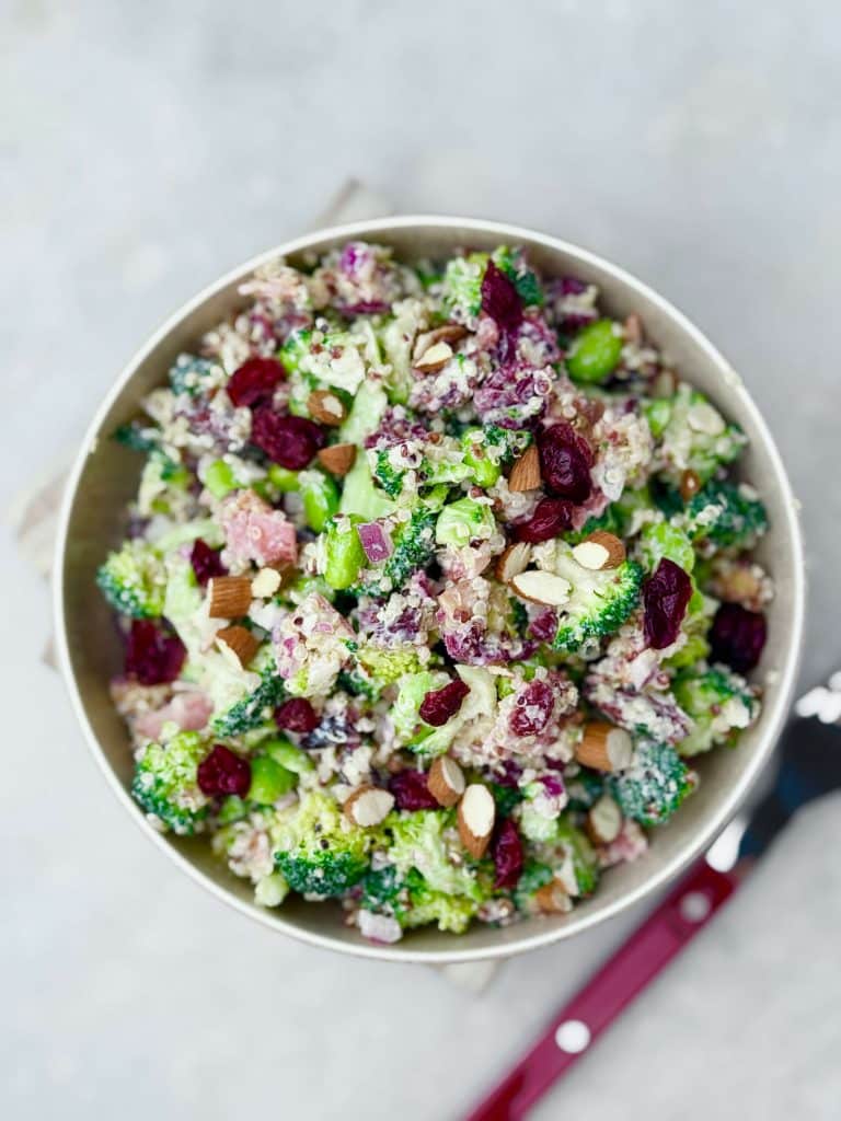 Top-down view of high protein broccoli salad tossed with dressing, featuring visible broccoli florets, bacon, almonds, edamame, and dried cranberries.