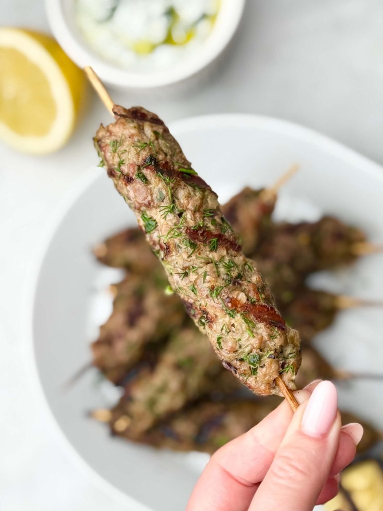 A single lamb kofta skewer held up close, showcasing grill marks and fresh herbs, with yogurt tzatziki in the background.