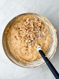 Chopped walnuts being folded into high protein banana bread muffin batter in a large bowl with a black spatula.