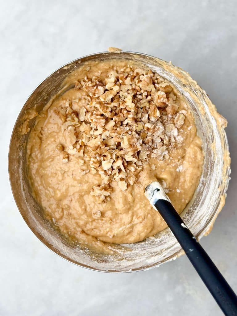Chopped walnuts being folded into high protein banana bread muffin batter in a large bowl with a black spatula.
